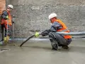 Dwóch budowlańców w kamizelkach odblaskowych wylewa pianobeton przez wąż na podłogę.