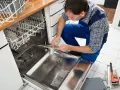 Fachowiec w niebieskim kombinezonie wykonuje podłączenie zmywarki, sprawdzając jej wnętrze. Obok leżą narzędzia.