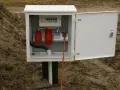 Skrzynka elektryczna na działce bez domu, gotowa do podłączenia prądu. Widoczne gniazda i zabezpieczenia.