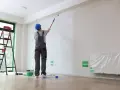 Malowanie ścian w mieszkaniu 50 m. Pracownik w kombinezonie maluje sufit wałkiem. Obok drabina, wiadro z farbą i folia ochronna.