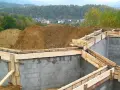 Fundamenty domu w budowie, zbrojenie i szalunki gotowe do zalania betonem. W tle widać kopiec ziemi i zabudowania. Pozwolenie na budowę zostało uzyskane.