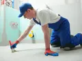 Budowlaniec w niebieskim kombinezonie i czapce nakłada pędzlem preparat betonit na podłogę w łazience.