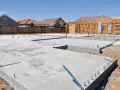 Fundamenty domu w budowie, betonowe płyty i zbrojenia. W tle szkielet drewniany domu, jakby chudziak czekał na dalsze prace.