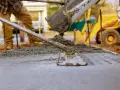 Robotnicy wylewają beton z miksokreta, wyrównując go łopatą. W tle żółta koparka.