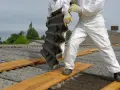 Osoby w białych kombinezonach usuwają z dachu eternit. Faliste płyty są demontowane z dachu pokrytego eternitem.