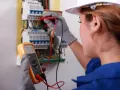 Kobieta w białym kasku i niebieskim kombinezonie wykonuje przegląd instalacji elektrycznej, używając multimetru.