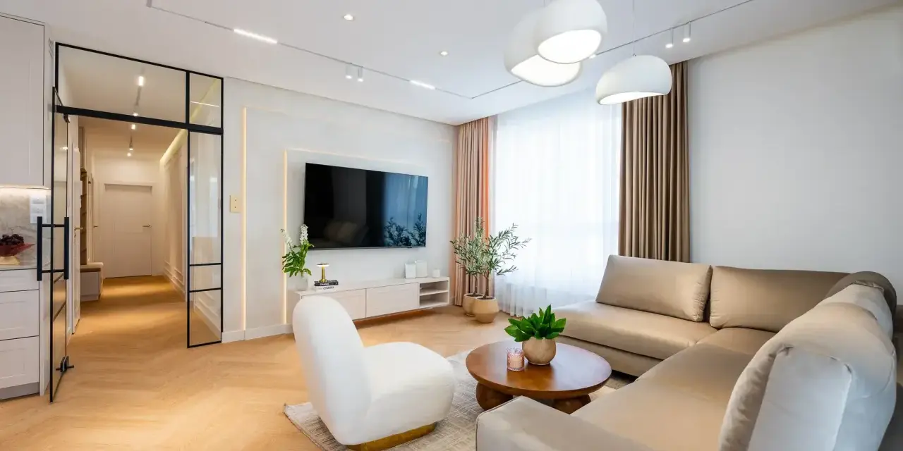 Nowoczesny salon z beżową kanapą, białym fotelem i drewnianą podłogą.