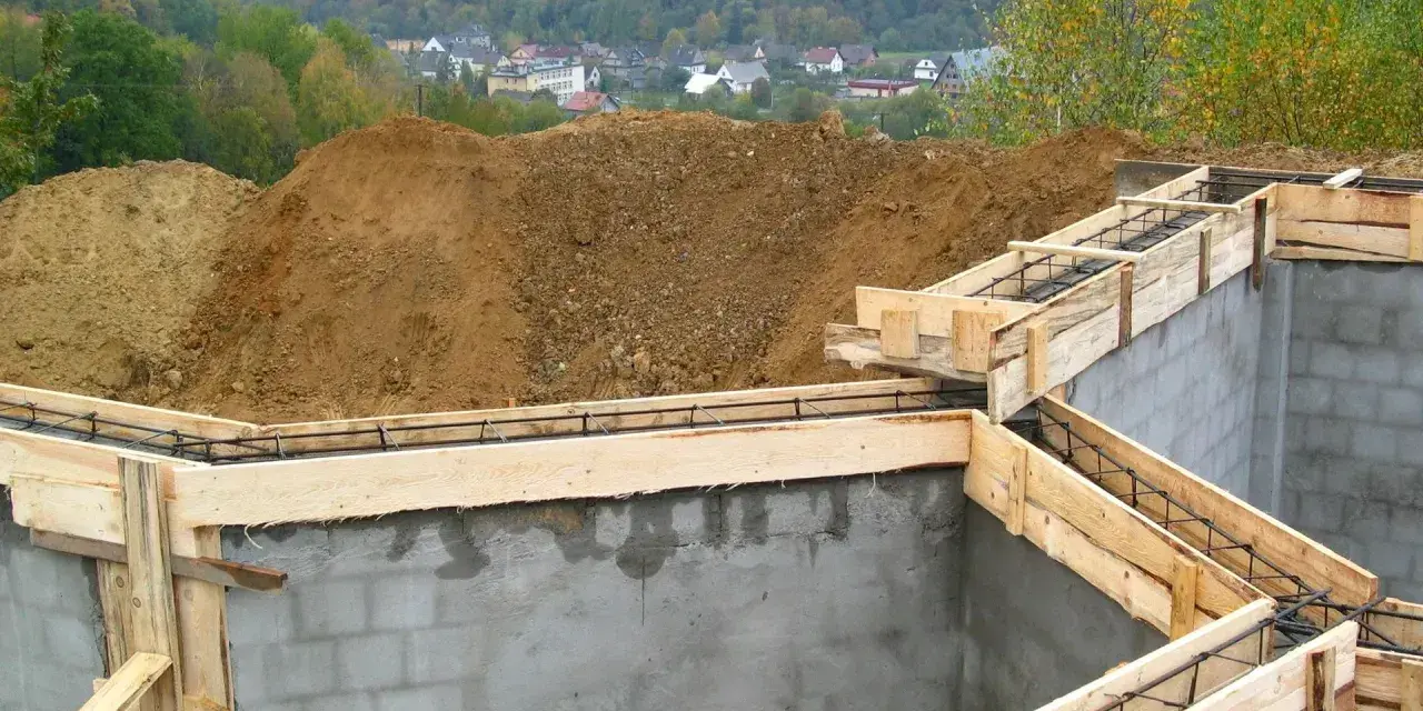 Fundamenty domu w budowie, zbrojenie i szalunki gotowe do zalania betonem. W tle widać kopiec ziemi i zabudowania. Pozwolenie na budowę zostało uzyskane.