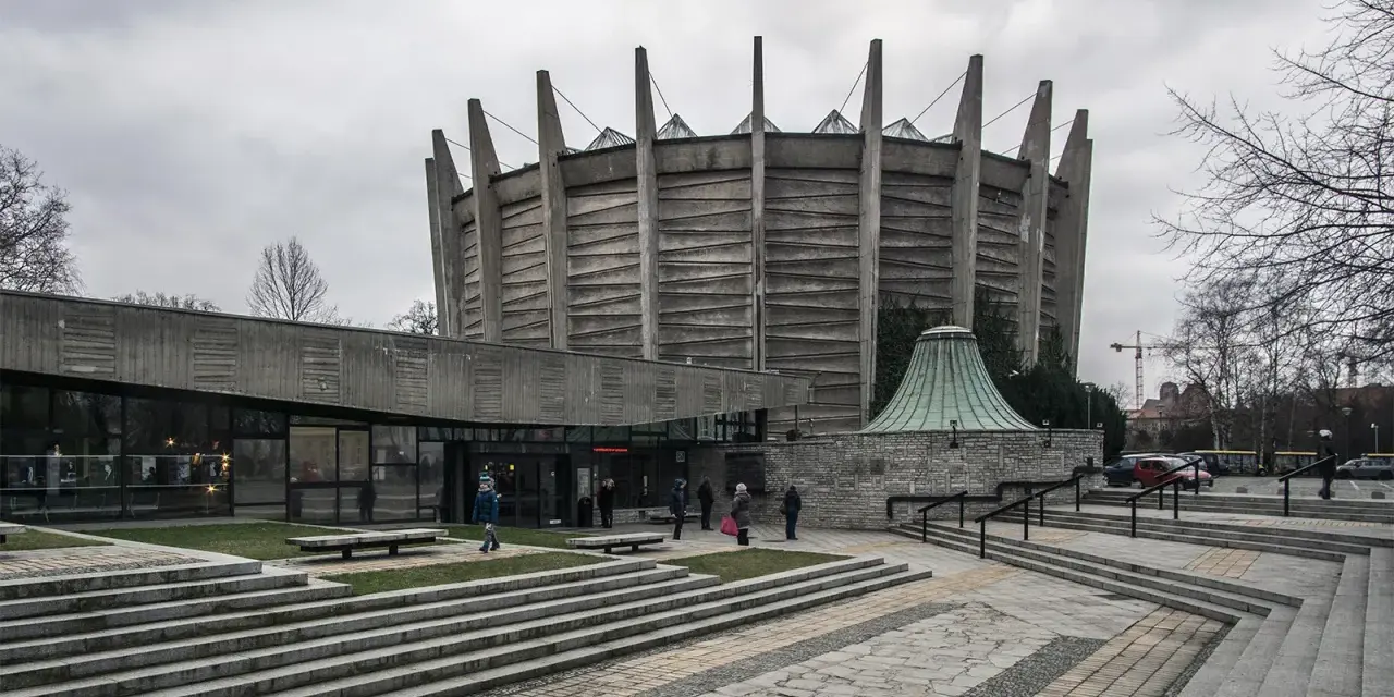 Monumentalny budynek o charakterze brutalizmu z betonowymi żebrami i szklanym wejściem. Schody prowadzą do wejścia, a wokół zielone trawniki.