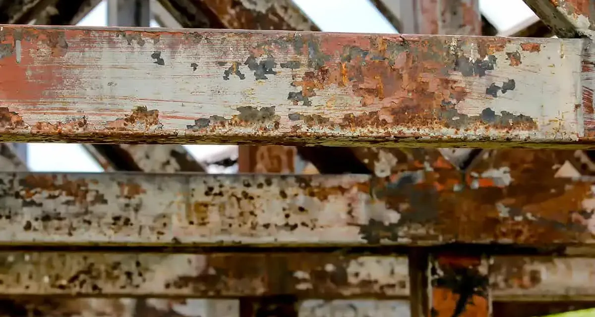 Metalowe belki konstrukcji z widoczną łuszczącą się farbą i rdzą. Postępująca korozja niszczy stal, tworząc rdzawy nalot i ubytki.