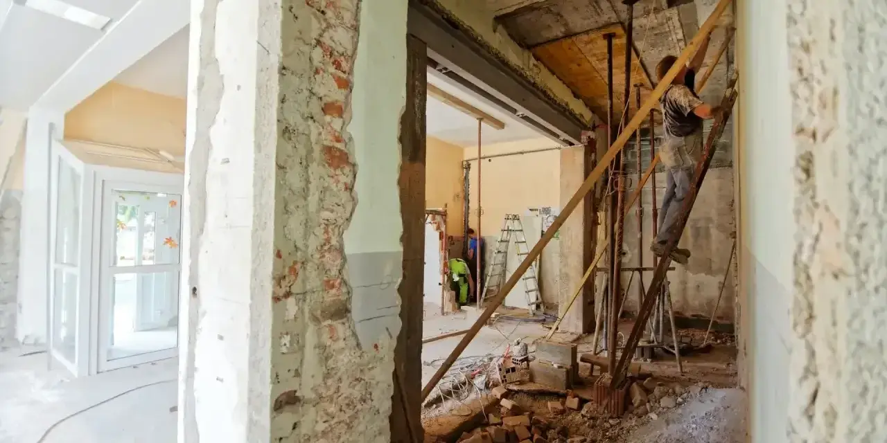 Trwa remont starego domu. Widać zburzone ściany, gruz i robotników pracujących przy konstrukcji.