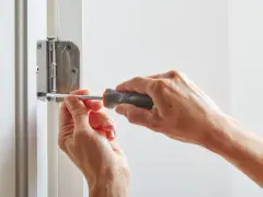 Dwie ręce przykręcają śrubę zawiasu, pokazując, jak wyregulować drzwi wejściowe.