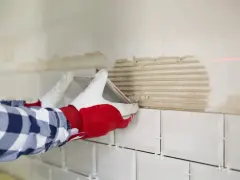 Ręce w rękawiczkach kładą białe płytki na ścianie z zaprawą.