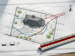 Plan zagospodarowania terenu z domem, drzewami i zaznaczonymi instalacjami. Linijka geodezyjna podkreśla skalę projektu.