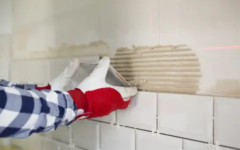 Ręce w rękawiczkach kładą białe płytki na ścianie z zaprawą.