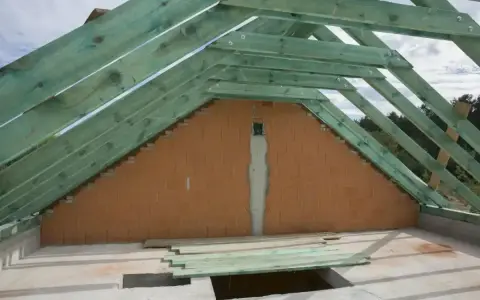 Widok na konstrukcję dachu i ścianę szczytową z cegły. Zielone drewniane krokwie tworzą szkielet dachu, a styk ściany szczytowej z dachem jest widoczny.