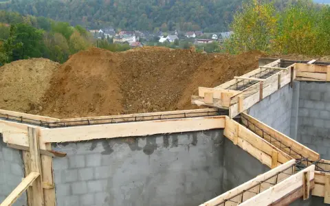 Fundamenty domu w budowie, zbrojenie i szalunki gotowe do zalania betonem. W tle widać kopiec ziemi i zabudowania. Pozwolenie na budowę zostało uzyskane.