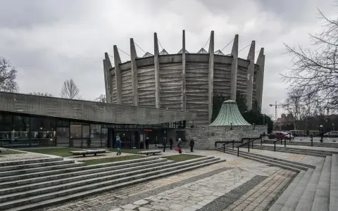 Monumentalny budynek o charakterze brutalizmu z betonowymi żebrami i szklanym wejściem. Schody prowadzą do wejścia, a wokół zielone trawniki.