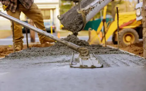 Robotnicy wylewają beton z miksokreta, wyrównując go łopatą. W tle żółta koparka.