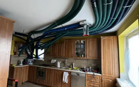 Instalacja rekuperacji w starym domu. W kuchni nad szafkami sufitowymi biegnie gęsta plątanina zielonych i niebieskich rur wentylacyjnych.