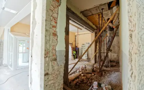 Trwa remont starego domu. Widać zburzone ściany, gruz i robotników pracujących przy konstrukcji.