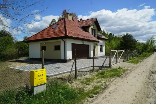 Nowy dom z garażem i żółtą skrzynką gazową. Brama i płot wyznaczają służebność drogi dojazdowej.