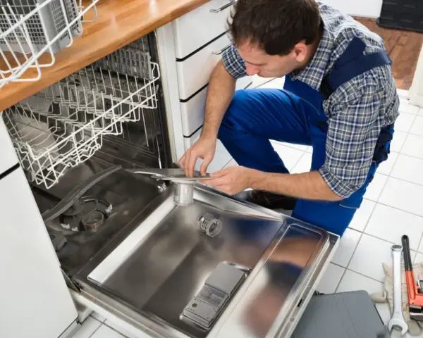 Fachowiec w niebieskim kombinezonie wykonuje podłączenie zmywarki, sprawdzając jej wnętrze. Obok leżą narzędzia.