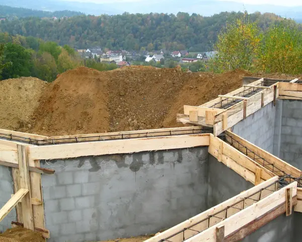 Fundamenty domu w budowie, zbrojenie i szalunki gotowe do zalania betonem. W tle widać kopiec ziemi i zabudowania. Pozwolenie na budowę zostało uzyskane.
