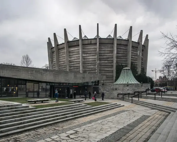 Monumentalny budynek o charakterze brutalizmu z betonowymi żebrami i szklanym wejściem. Schody prowadzą do wejścia, a wokół zielone trawniki.
