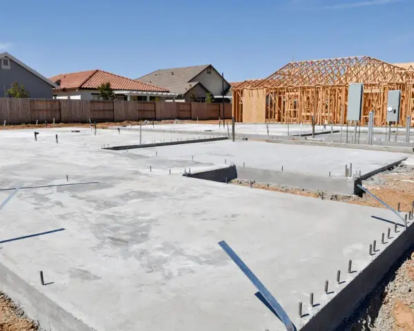 Fundamenty domu w budowie, betonowe płyty i zbrojenia. W tle szkielet drewniany domu, jakby chudziak czekał na dalsze prace.