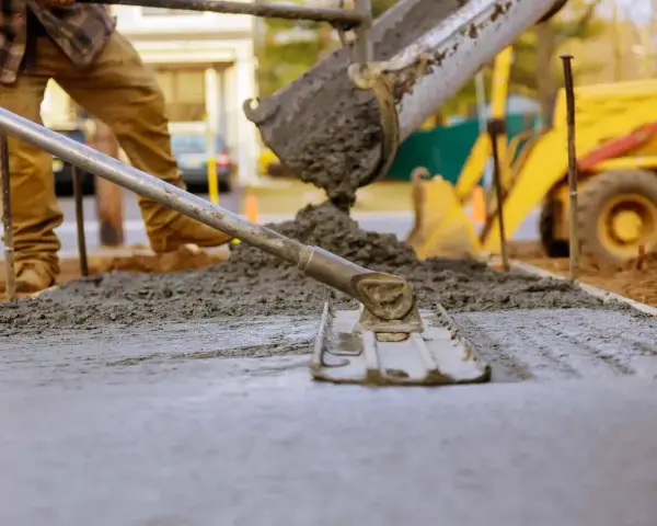 Robotnicy wylewają beton z miksokreta, wyrównując go łopatą. W tle żółta koparka.