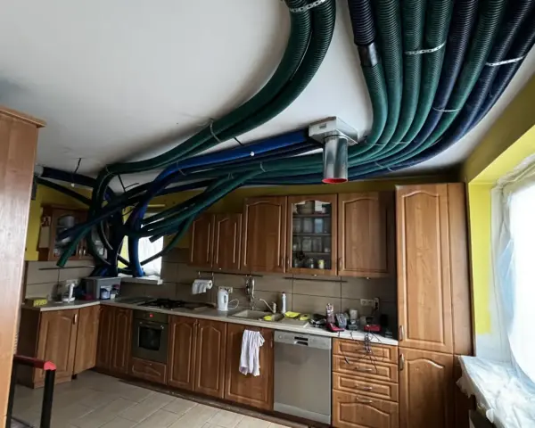 Instalacja rekuperacji w starym domu. W kuchni nad szafkami sufitowymi biegnie gęsta plątanina zielonych i niebieskich rur wentylacyjnych.