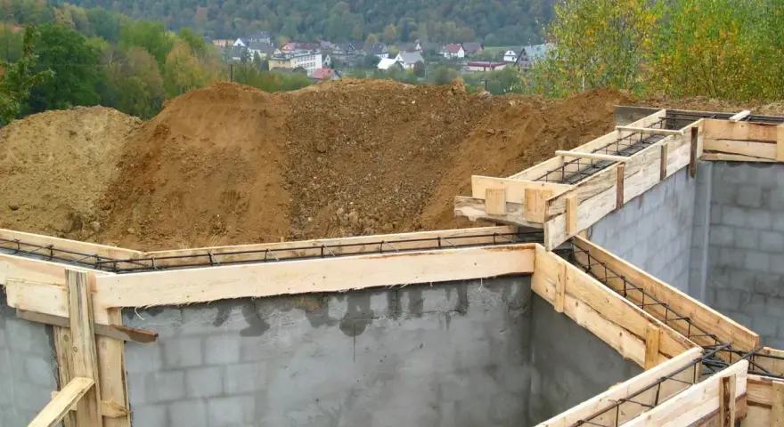 Fundamenty domu w budowie, zbrojenie i szalunki gotowe do zalania betonem. W tle widać kopiec ziemi i zabudowania. Pozwolenie na budowę zostało uzyskane.
