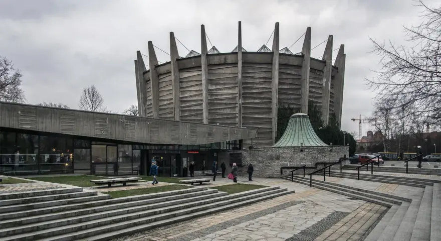 Monumentalny budynek o charakterze brutalizmu z betonowymi żebrami i szklanym wejściem. Schody prowadzą do wejścia, a wokół zielone trawniki.