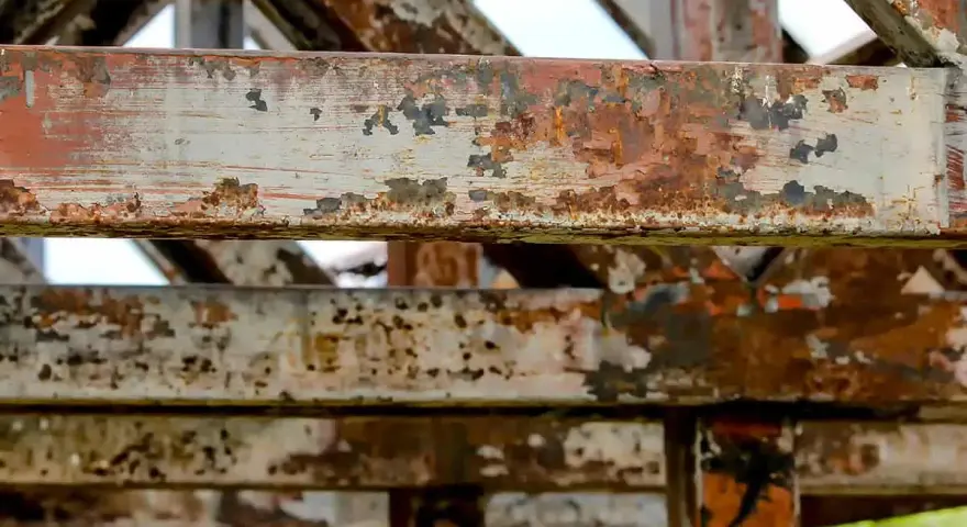 Metalowe belki konstrukcji z widoczną łuszczącą się farbą i rdzą. Postępująca korozja niszczy stal, tworząc rdzawy nalot i ubytki.