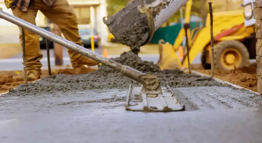 Robotnicy wylewają beton z miksokreta, wyrównując go łopatą. W tle żółta koparka.
