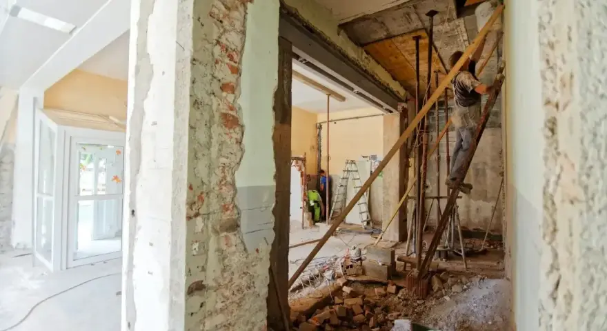 Trwa remont starego domu. Widać zburzone ściany, gruz i robotników pracujących przy konstrukcji.