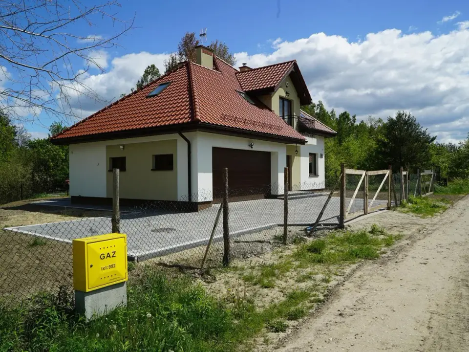 Nowy dom z garażem i żółtą skrzynką gazową. Brama i płot wyznaczają służebność drogi dojazdowej.