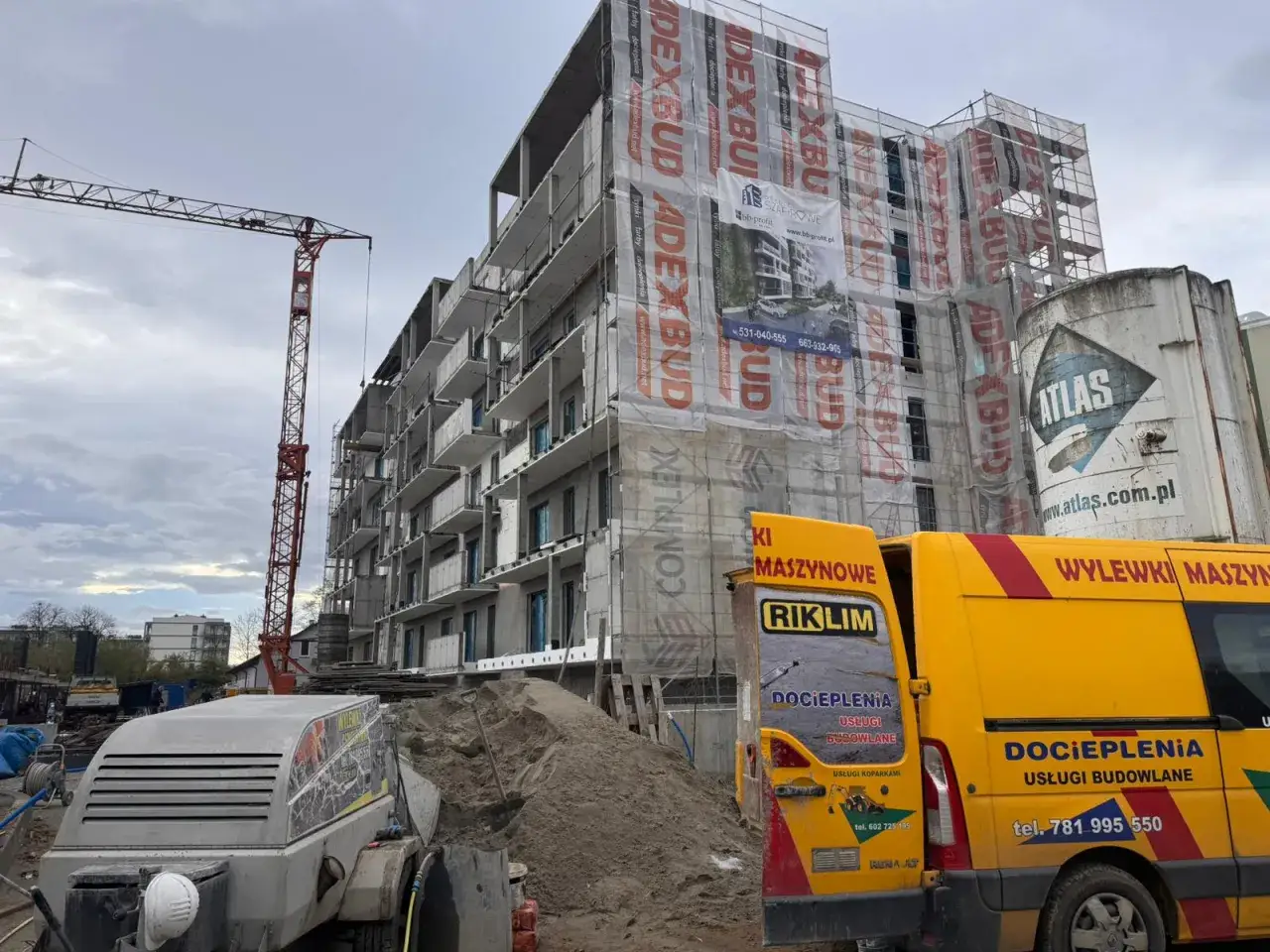 Plac budowy z dźwigiem, niedokończonym budynkiem i żółtymi samochodami. Widać miksokret do wylewek.