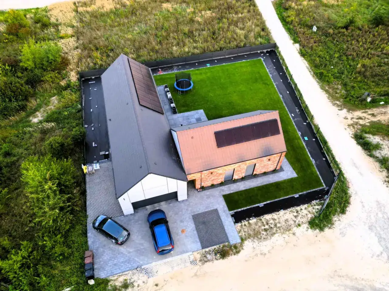 Nowy dom pod klucz z panelami słonecznymi, zadbanym ogrodem i trampoliną. Dwa samochody zaparkowane przed garażem.