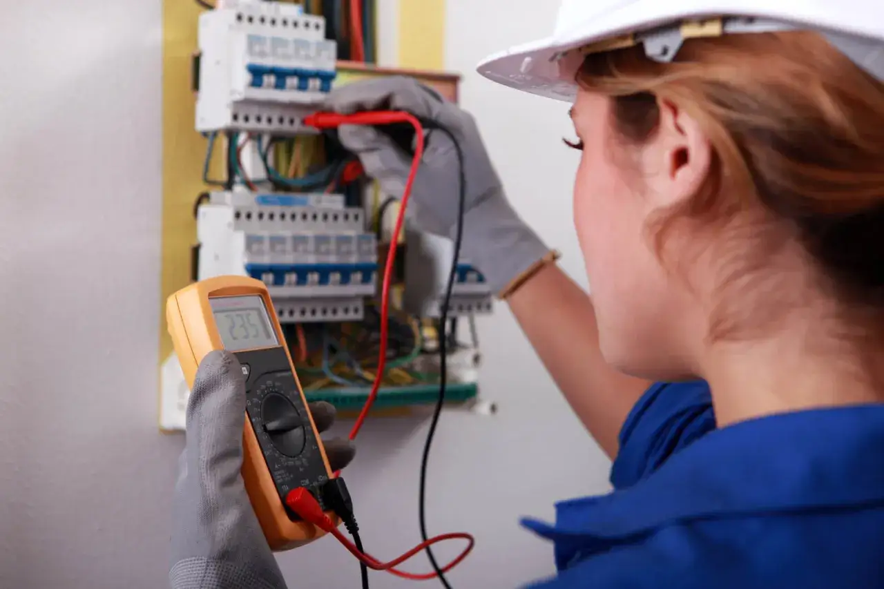 Elektryk w białym kasku przeprowadza przegląd instalacji elektrycznej, używając multimetru do pomiaru napięcia.