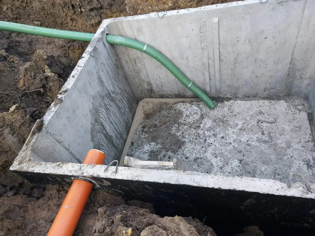 Montaż szamba betonowego 10m3. Widoczny betonowy zbiornik w wykopie, z podłączonymi rurami.